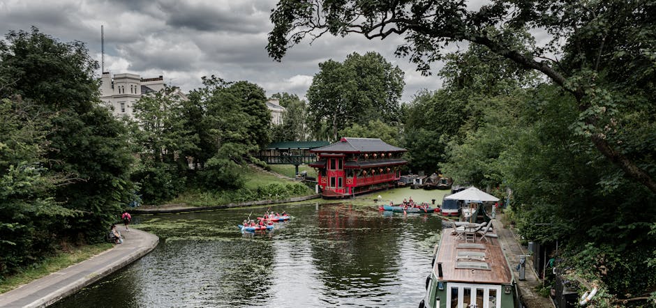 A wide view of the Regent's Canal in Little Venice, London, showing calm water with several small boats and kayaks being paddled by people in life jackets. On the right side, a narrow towpath features a small docked houseboat with a weathered wooden roof and metal chimneys. To the left, a stone walkway runs alongside the canal, with a person walking and another sitting on a bench. Dense green trees and foliage line the edges of the canal, leading to a historic red and black pagoda-style building with traditional architecture, situated near the water's edge, partly shaded by trees. In the background, large white residential buildings are partially visible through the treetops under a cloudy sky, creating a peaceful scene associated with house removals and relocation logistics in the Paddington area, as highlighted by Paddington Man and Van's services.