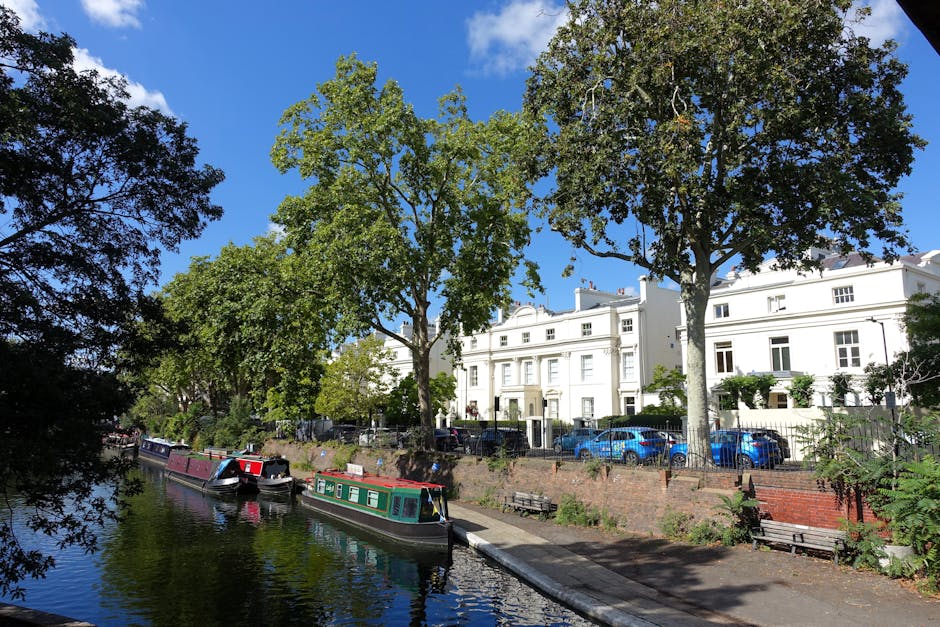 A daytime scene along a canal in Little Venice, featuring a narrowboat moored parallel to the water's edge. In the foreground, the canal reflects the surrounding environment, with calm water and some trees casting shadows on the banks. Alongside the canal, there is a paved walkway with a few benches, and a low brick wall separates the path from residential buildings behind it. The buildings are white, multi-story Victorian-style houses with large windows, some with balconies. Parked cars, mostly blue, are visible along the street in front of the houses. Tall, leafy trees with green foliage line the street, partially obscuring parts of the buildings and providing shade. In the background, a clear blue sky with a few clouds can be seen. This scene illustrates the typical setting for house removals and furniture transport operations in the area, with Paddington Man and Van specialising in local relocation and moving services, including loading and packing at the property and careful handling of household items for a smooth home relocation process.